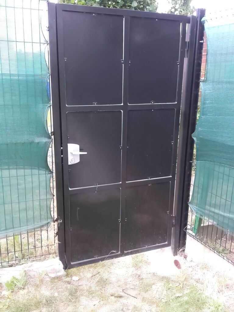 Portillon en métal noir avec serrure de sécurité, installé dans une clôture avec des panneaux occultants verts.