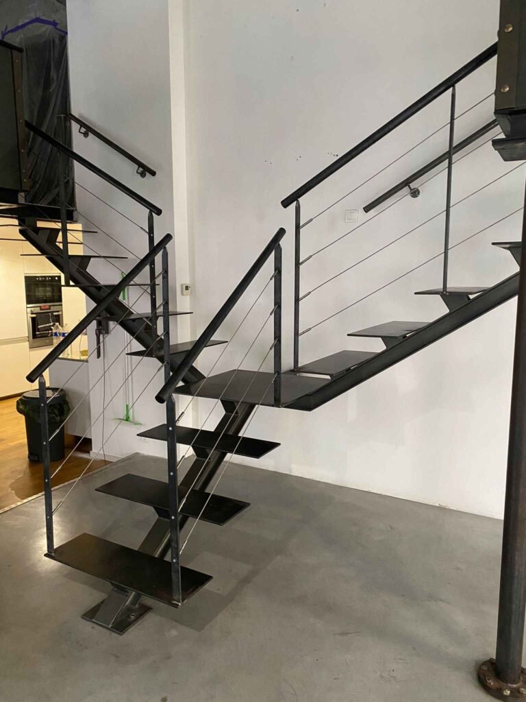 Un escalier intérieur moderne avec des marches en métal noir, une rampe en câbles métalliques et des montants noirs, menant à un étage supérieur dans un espace ouvert avec une cuisine visible.